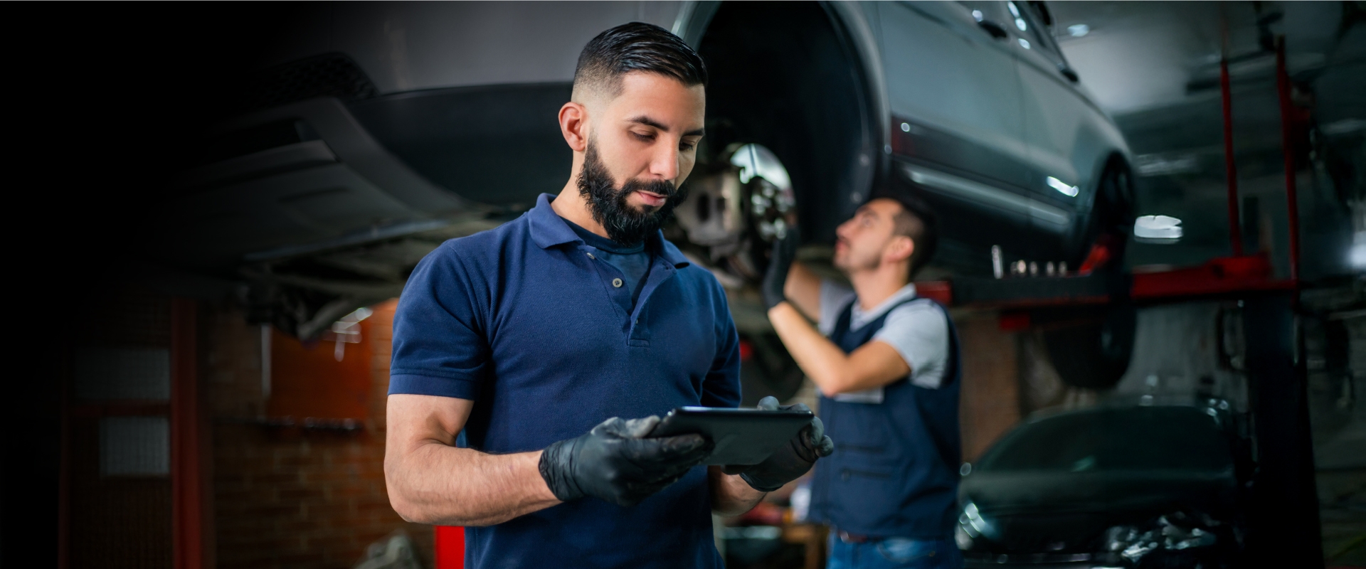 Mechanik samochodowy w warsztacie sprawdza dane na tablecie, podczas gdy drugi mechanik pracuje przy podniesionym aucie na podnośniku.
